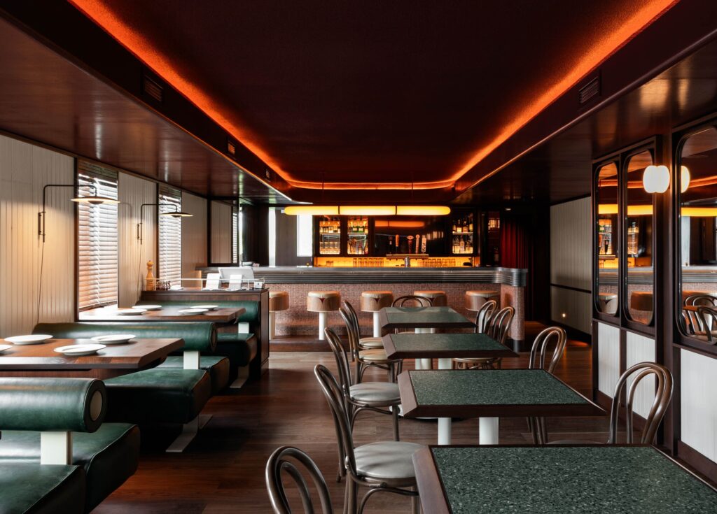 Interior view of North of Brooklyn Pizzeria showing green leather booths, walnut tables, and a long bar under warm ambient orange lighting.