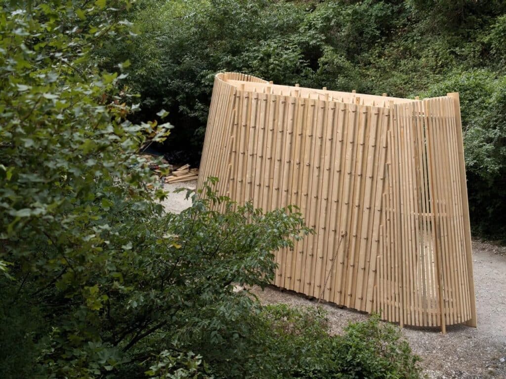 Elevated wide shot of the La Barca pavilion, a curved wooden structure located on a gravel path surrounded by dense green vegetation.