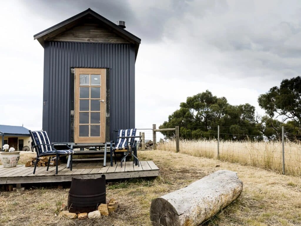 Front entrance of a modern tiny house on wheels with a wooden door and small outdoor deck in a farm setting.