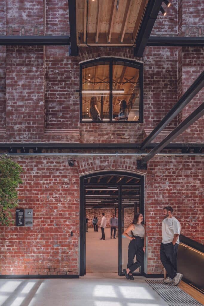 People socializing in an open-plan heritage office space with exposed brick walls, timber ceilings, and arched windows at Younghusband Melbourne.