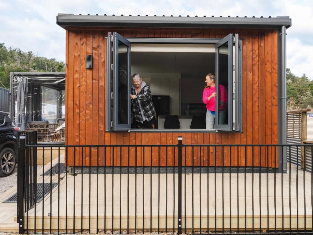 Residents looking out of a large black-framed bifold window of the Bespoke Base tiny home with Redwood siding.