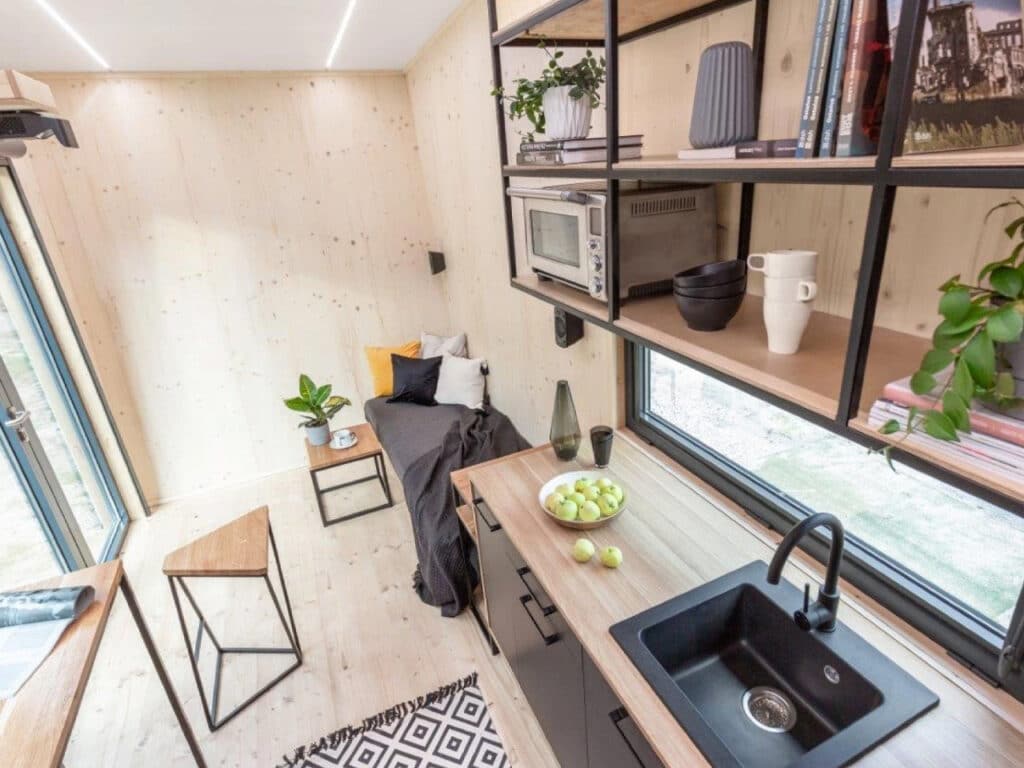 Modern kitchenette in a tiny house with a black sink, wooden countertops, and open industrial shelving overlooking a seating area.