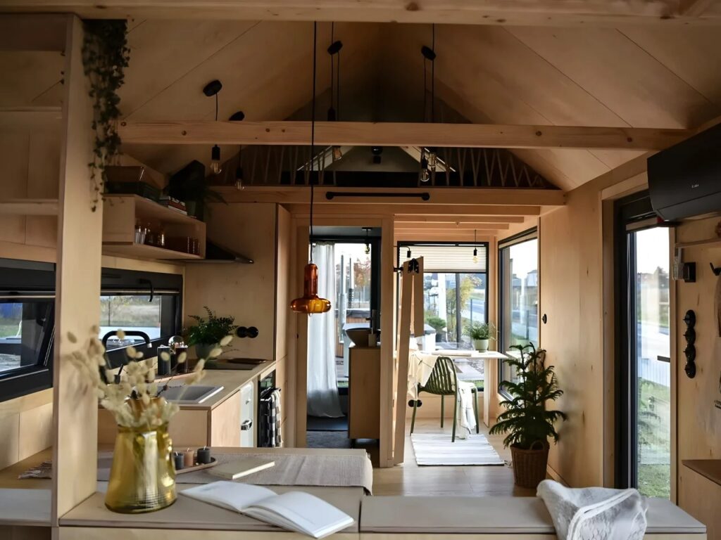 Wide interior view of Julia tiny home showing the open-plan layout, dining area, and high-pitched wooden ceiling with hanging lights.