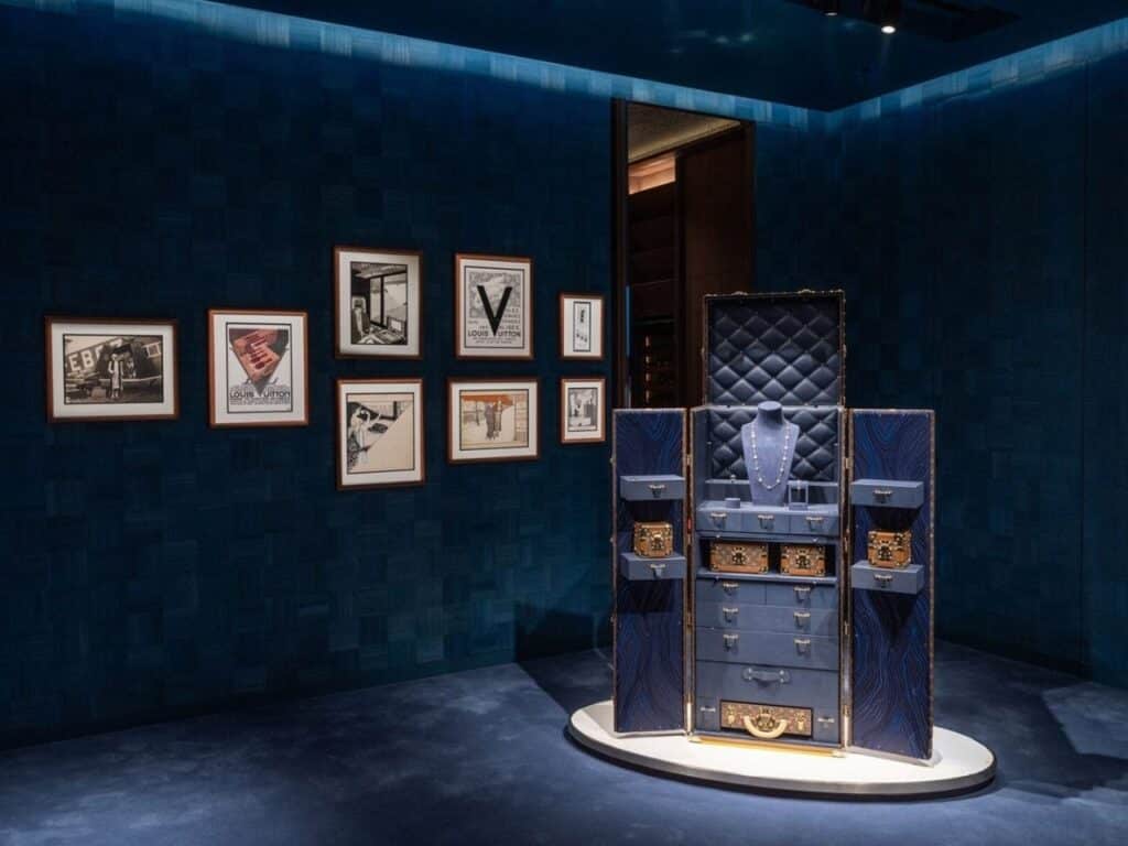 Artistic display of a classic Louis Vuitton trunk against a dark blue textured wall with framed heritage sketches at the Beijing Maison.
