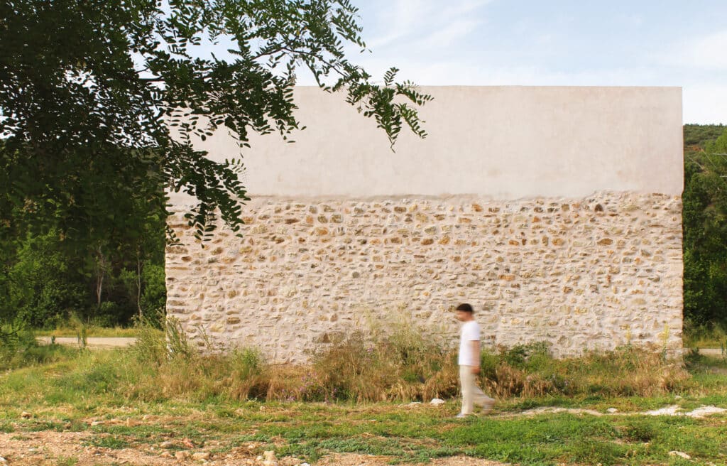 Person walking past a long, textured stone and rammed earth wall with a minimalist white facade above it.