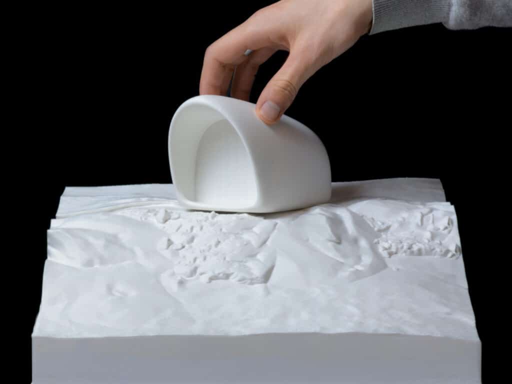A hand placing a 3D architectural model of the AltiHut on a topographic base.