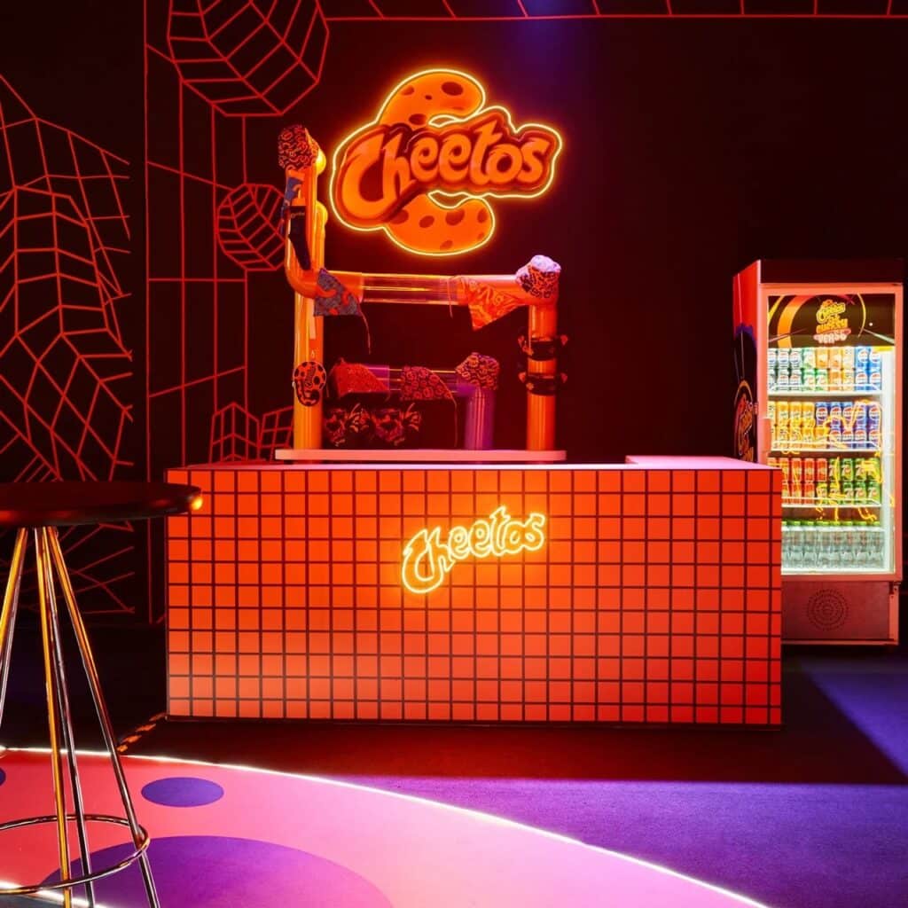 Modern snack bar with a glowing neon Cheetos logo, grid-patterned counter, and a beverage cooler in a dark-themed interactive space.