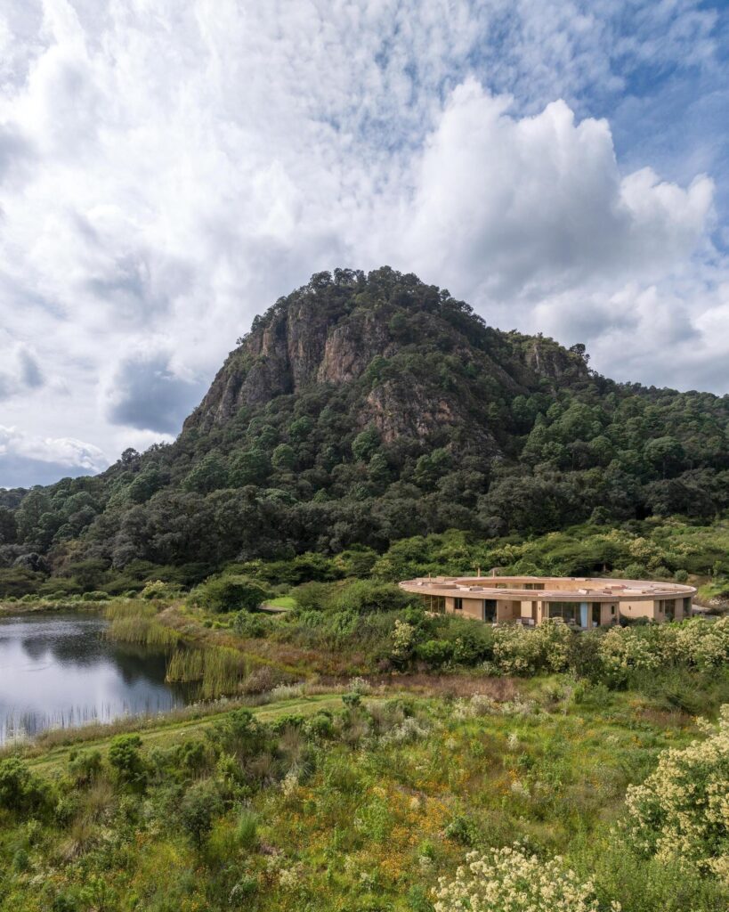 Aerial view of the circular Degrees house nestled in a green valley next to a lake and a large mountain.