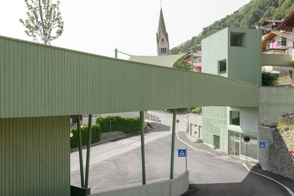 A green wooden pedestrian bridge spanning a public street, connecting two educational buildings in the mountain village of Barbiano.
