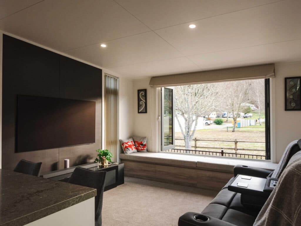 Interior living room of a tiny house with a large wall-mounted TV, gray sofa, and a long built-in window seat.