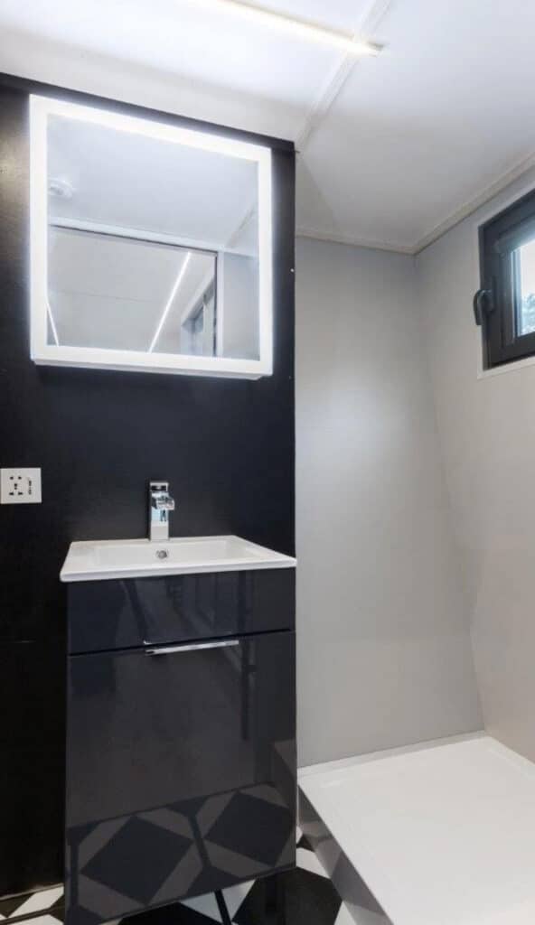 Minimalist tiny house bathroom featuring a black modern vanity, illuminated mirror, and a sleek walk-in shower.