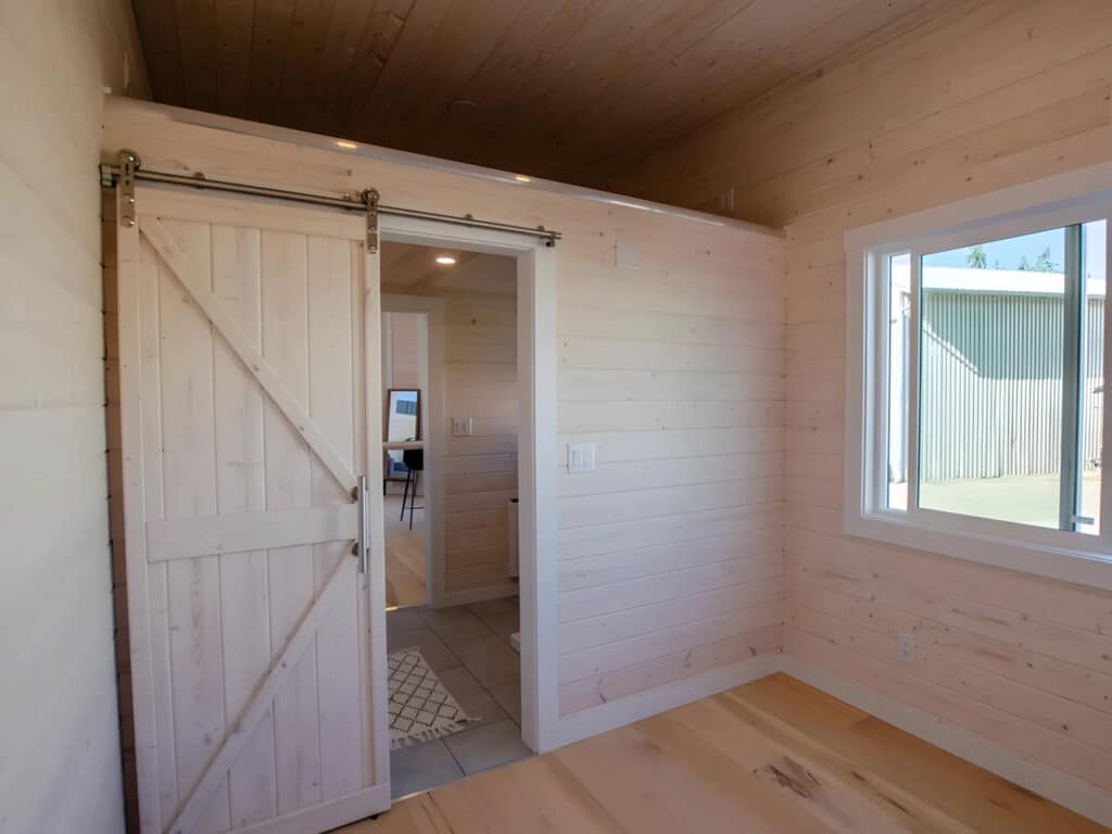 Interior bedroom of the Dove tiny home showing the ground-floor sleeping area and a white sliding barn door.