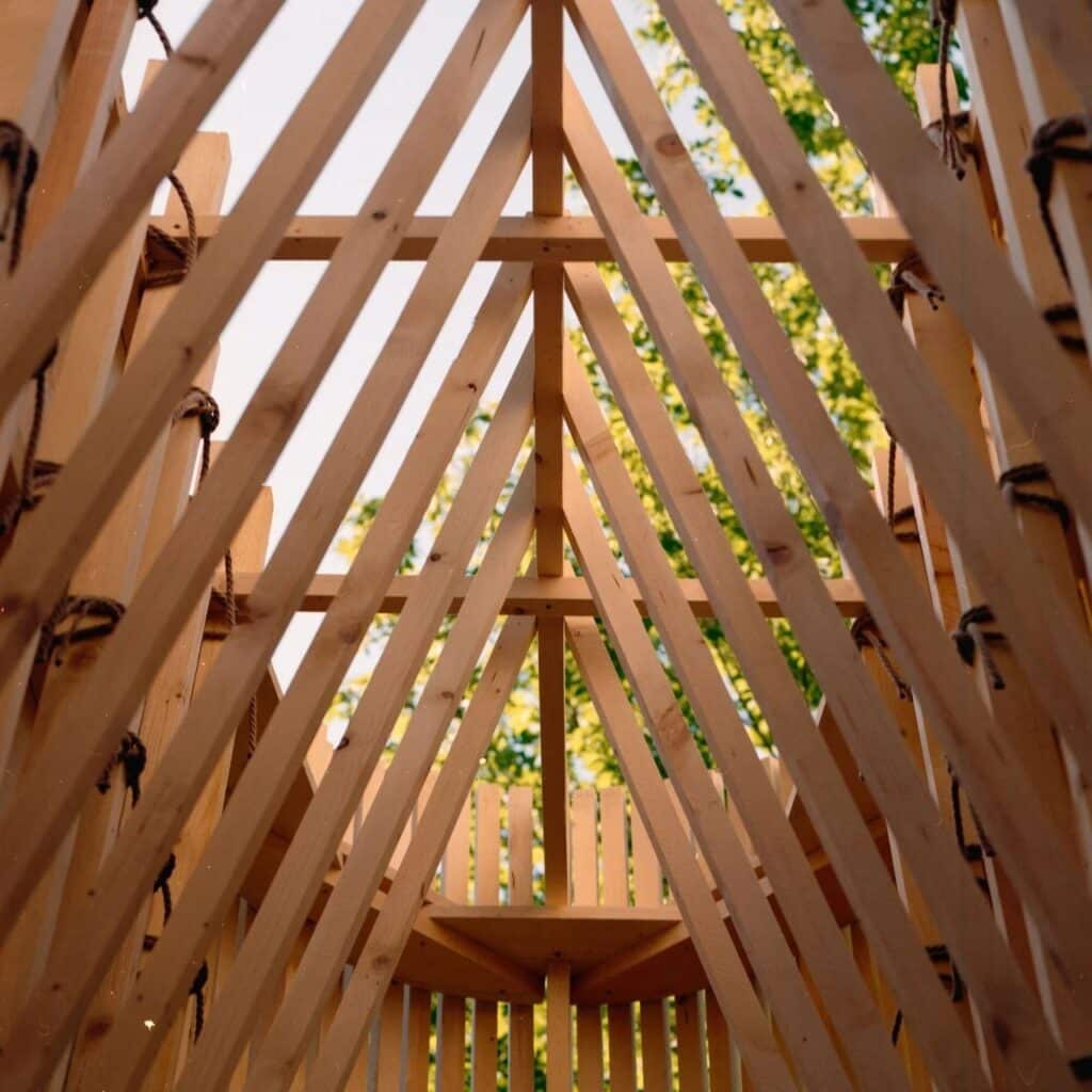 Low-angle interior view of La Barca wooden pavilion showing the geometric timber roof structure and rope bindings against a backdrop of green trees.