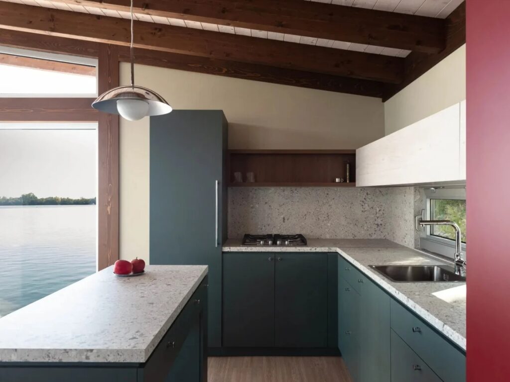 Minimalist kitchen area in the Sava River Cabin with teal cabinets, stone countertops, and a window reflecting the river.