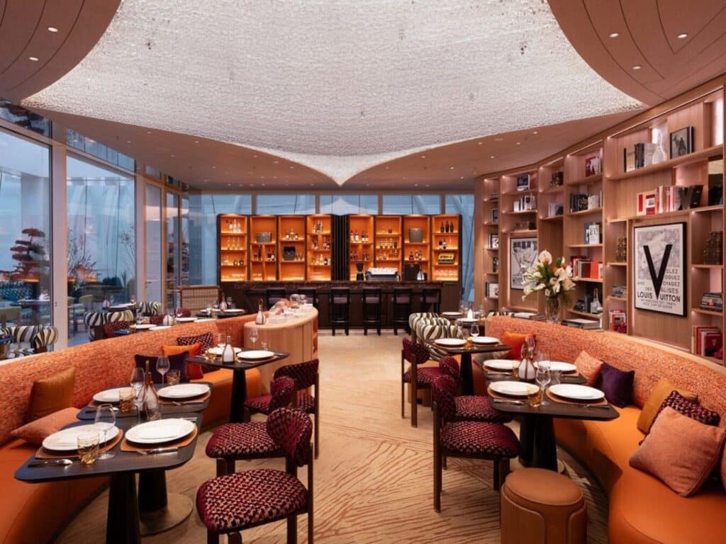 Spacious dining area of Le Caf&eacute; Louis Vuitton in Beijing with warm orange seating, textured ceiling, and views of the Sanlitun district through the glass skin.