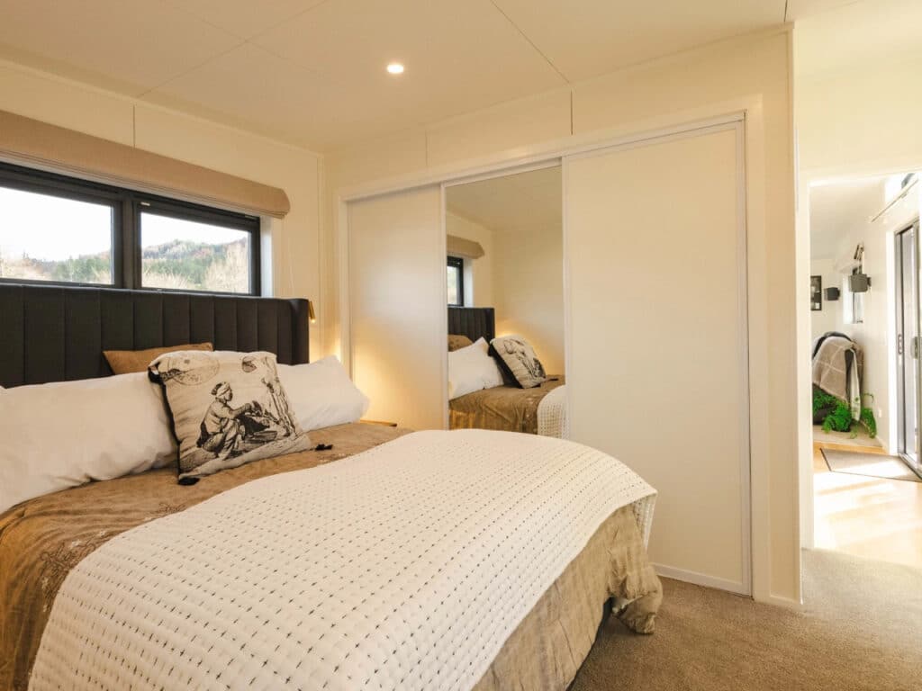 Modern bedroom in a tiny home with a large bed, built-in mirrored wardrobes, and a large window.