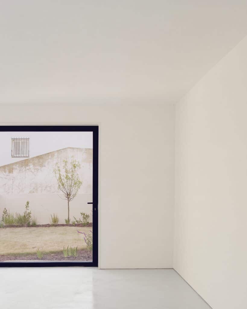 Interior view of a white minimalist room looking out through a large window to a garden.