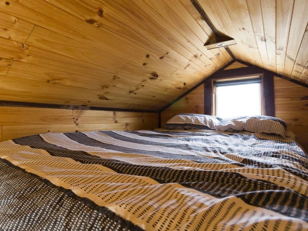 Cozy sleeping loft in a tiny house with a queen-sized bed under a pitched wooden ceiling.