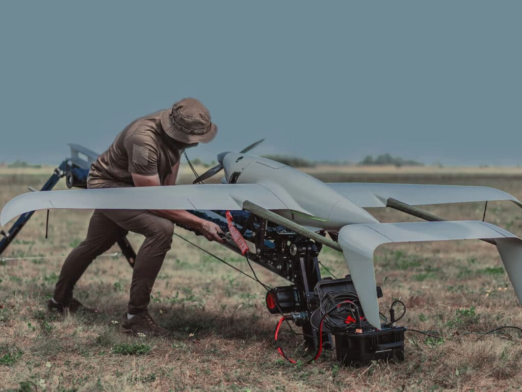 Military operator in a boonie hat adjusting the Raybird drone on its launch platform in a field.