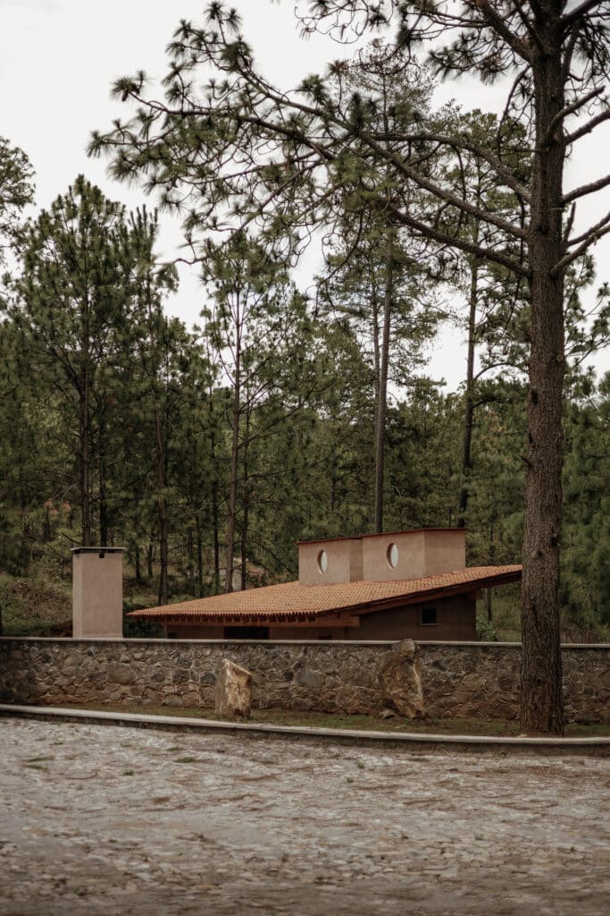 Low-angle view of the cabin's sloping roof and circular windows behind a traditional stone wall in the forest.