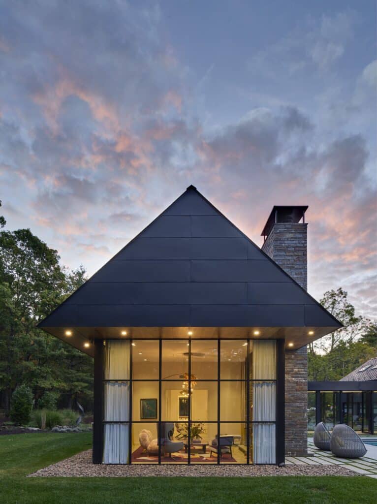 Night view of a gabled living room with large glass windows and a stone chimney at WACE House.