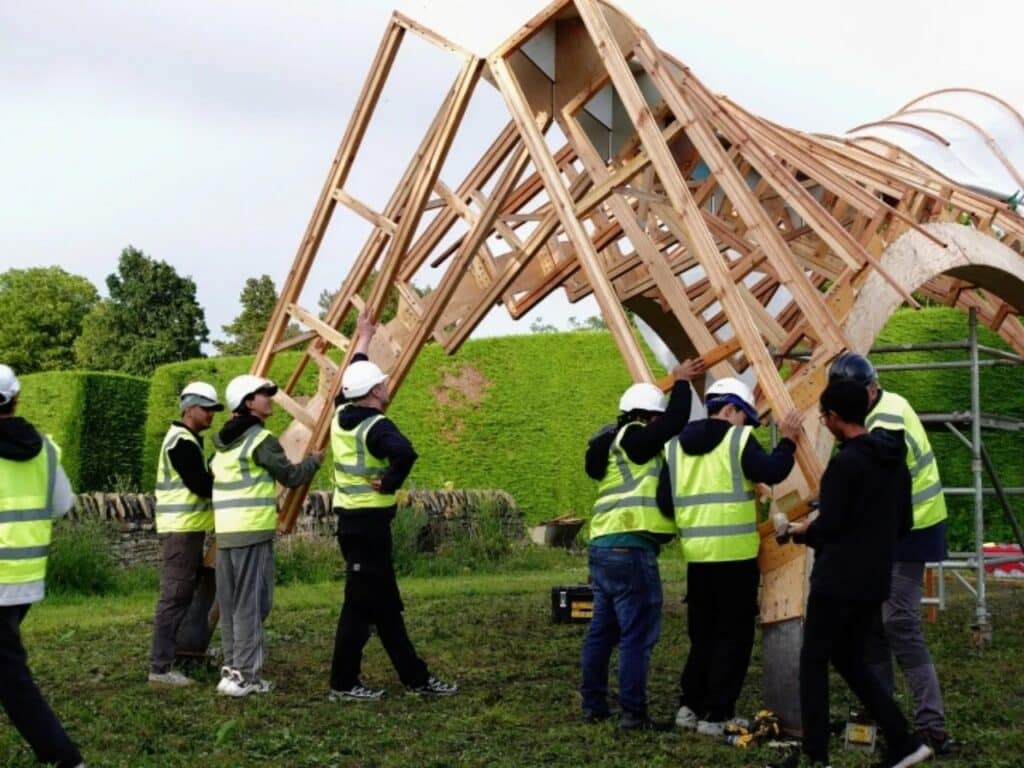Architecture students in high-visibility vests manually assembling the robotic-made timber structure of the Arkhive pavilion outdoors.