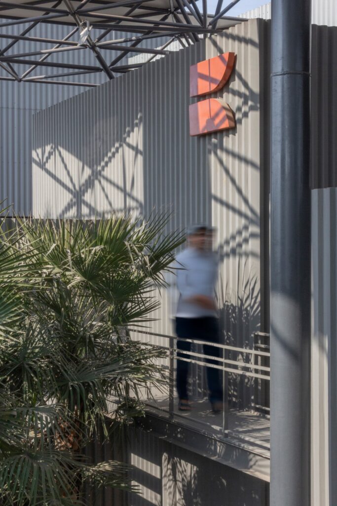 Close-up of the Karachi Steel orange logo on a grey corrugated wall with shadows from the space frame and a person walking on a bridge.