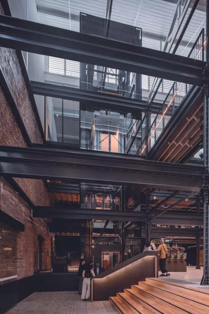 Modern glass elevator and black industrial steel beams integrated with original Victorian red brick walls and timber stairs in the Younghusband building.