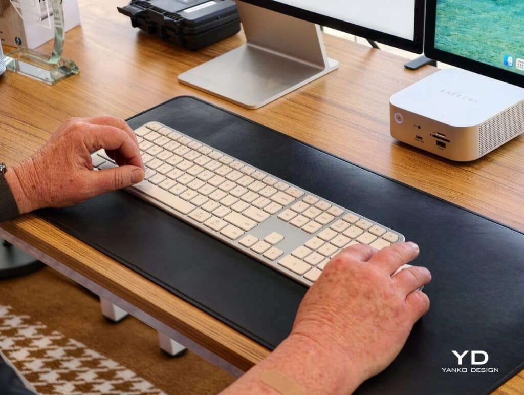Hands typing on a silver Satechi Slim EX3 full-size keyboard on a wooden desk with a leather desk mat and a Mac Studio.