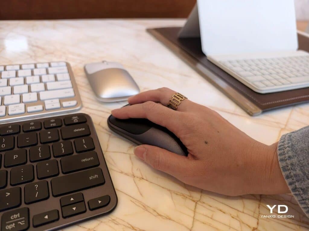 A person's hand using a space gray Satechi Slim EX wireless mouse on a marble desk with a keyboard and iPad in the background.