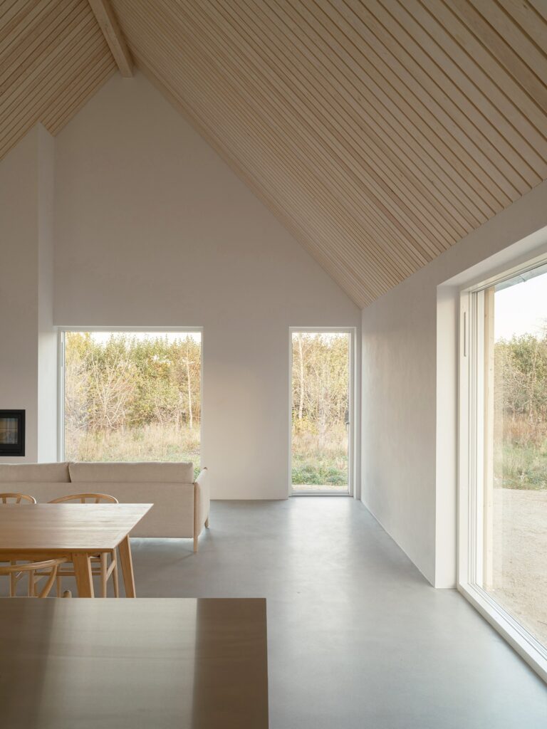 Interior view of Rorum House living area with double-height ceilings, pine wood cladding, and large windows facing the orchard.
