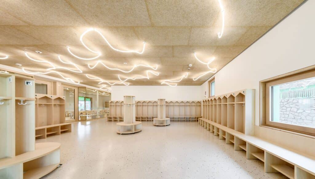 Minimalist wooden cloakroom for children in the Barbiano kindergarten with integrated cubbies and wavy overhead lighting.
