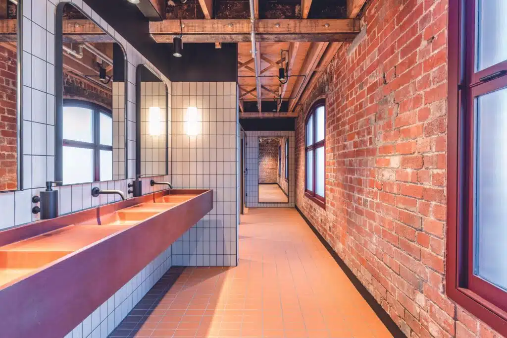 A renovated industrial-style bathroom featuring red brick walls, exposed timber ceiling beams, and a long terracotta-colored trough sink.