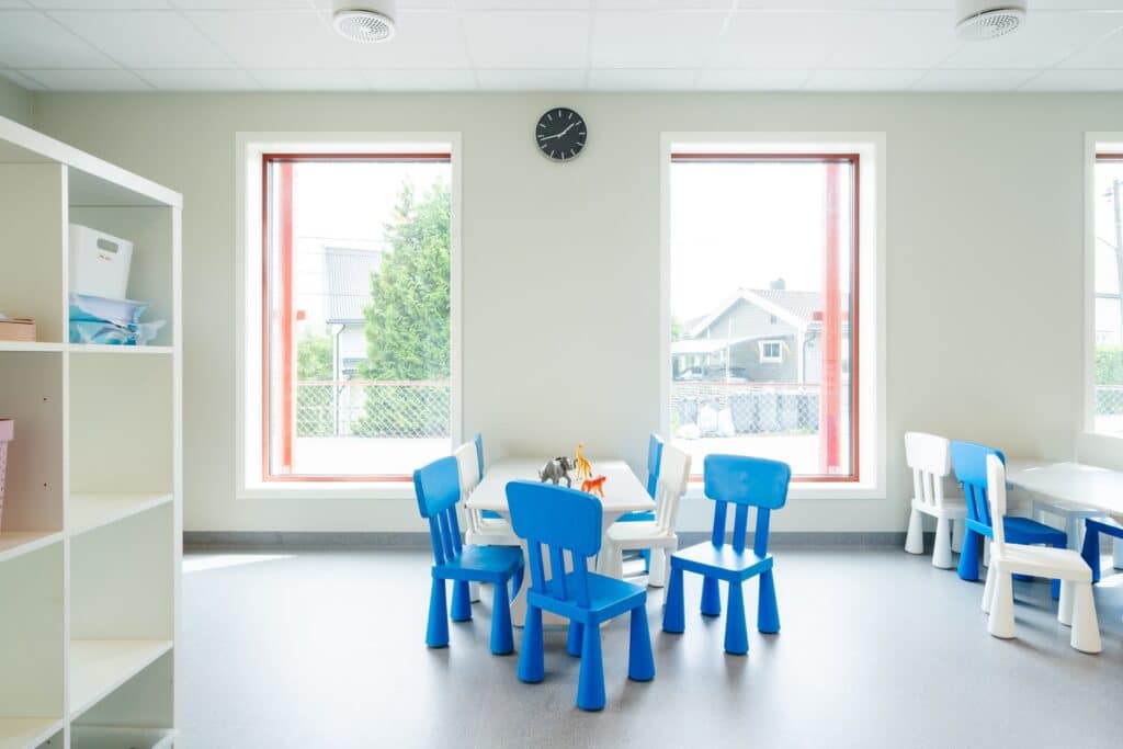 Interior of a playroom at Alvim Kindergarten featuring large low-silled windows, blue chairs, and a grey wall backdrop.