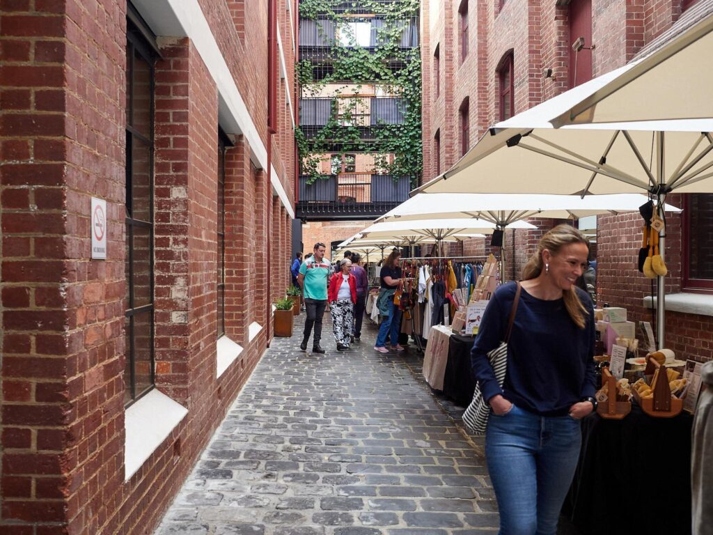 A vibrant outdoor pedestrian alleyway with bluestone paving between historic red brick buildings, featuring community market stalls under umbrellas.