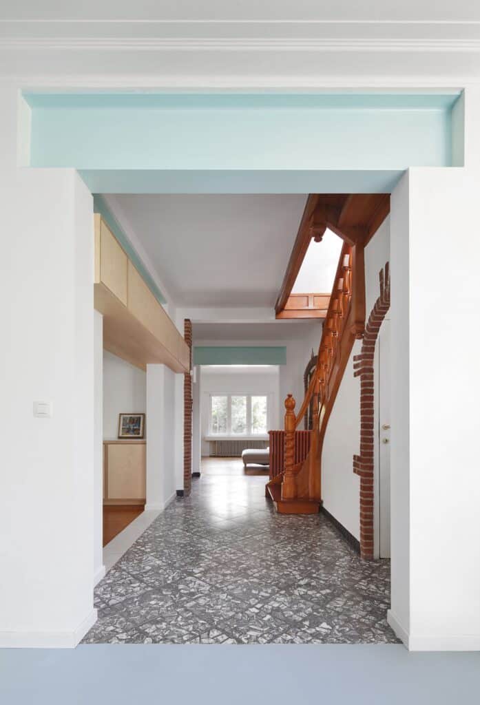 Interior hallway of a Brussels home showing a preserved classic wooden staircase and terrazzo flooring under a modern light-blue lintel.