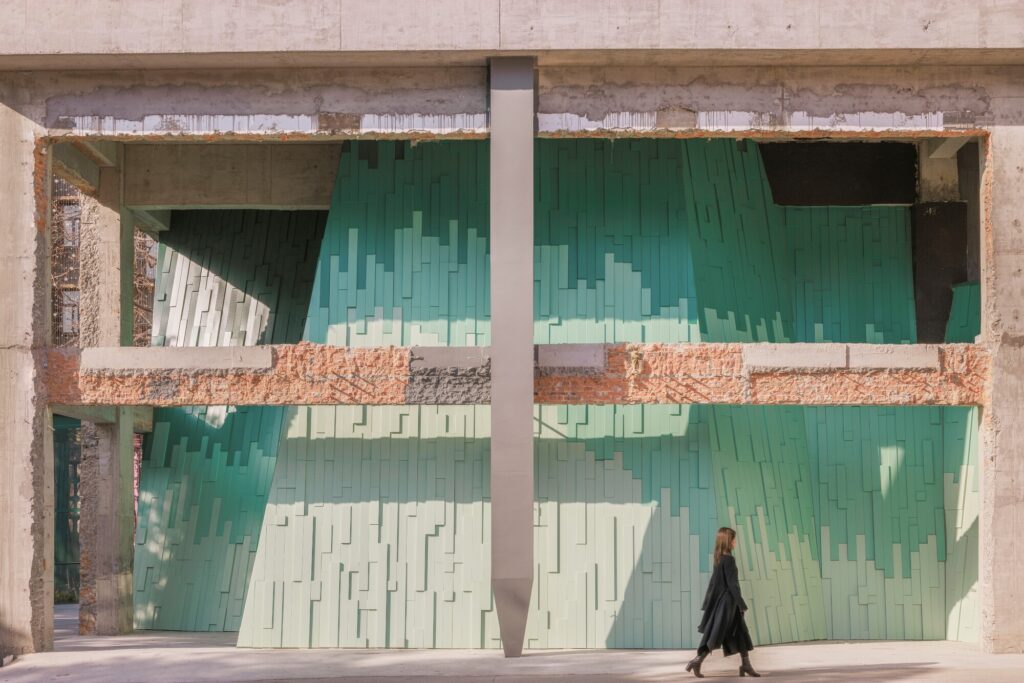 A wide view of the south elevation showing a person walking through the open industrial concrete frame that houses the green mountain-like facade.