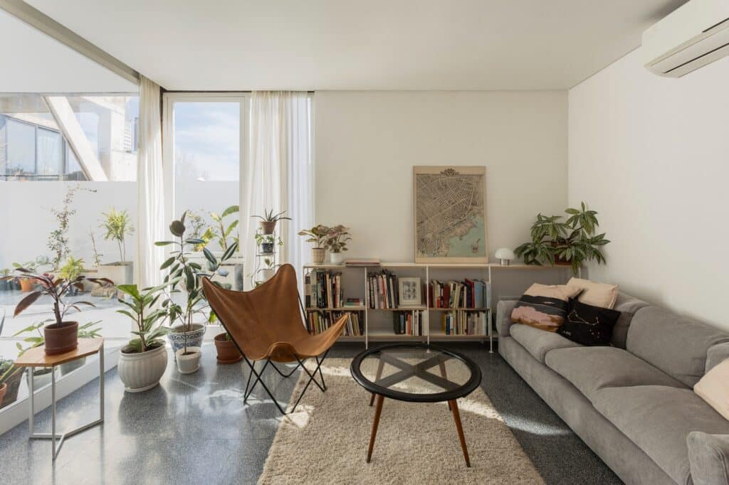 Modern living room interior with a grey sofa, butterfly chair, and large glass windows overlooking a sunlit patio.