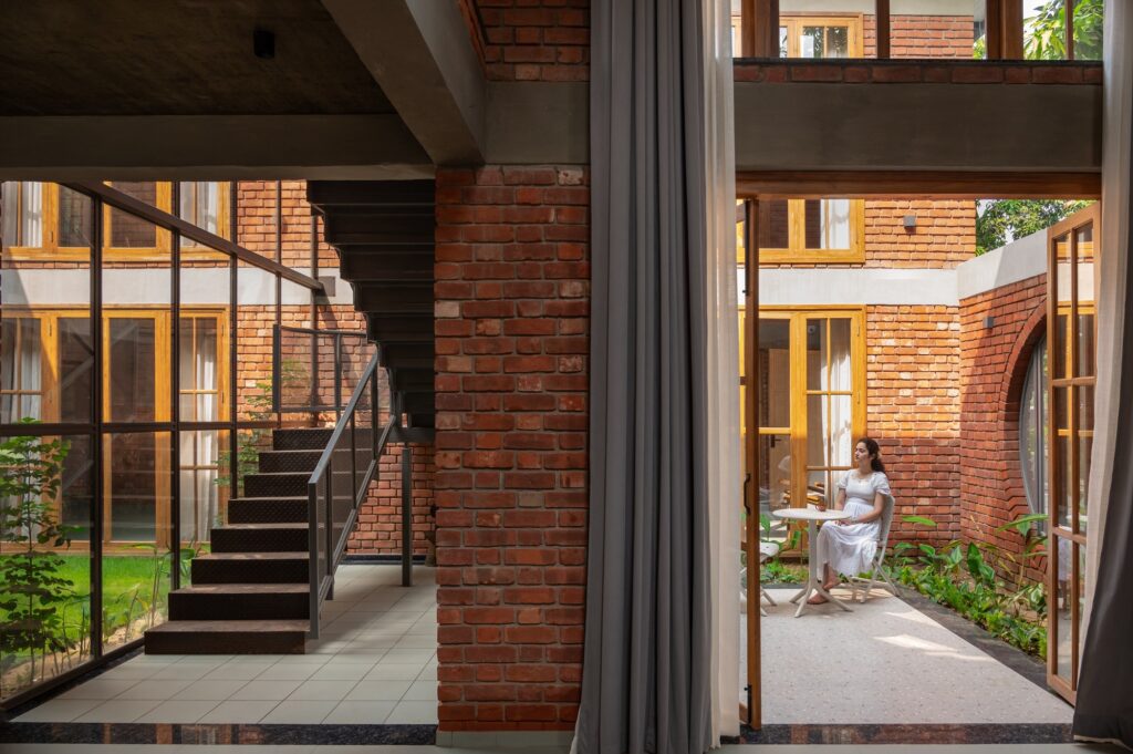 A woman sitting in a quiet courtyard with exposed brick walls, a circular architectural opening, and a metal staircase in the background.