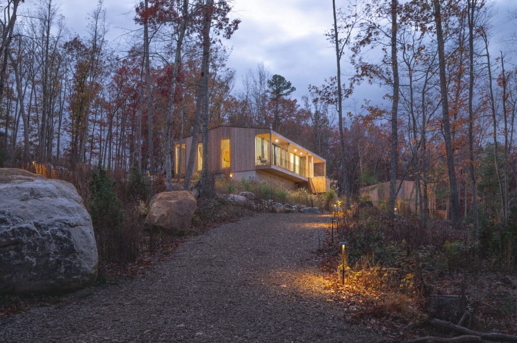Sera project villa illuminated at dusk, nestled in the forest with a gravel path leading to the glowing sustainable structure.