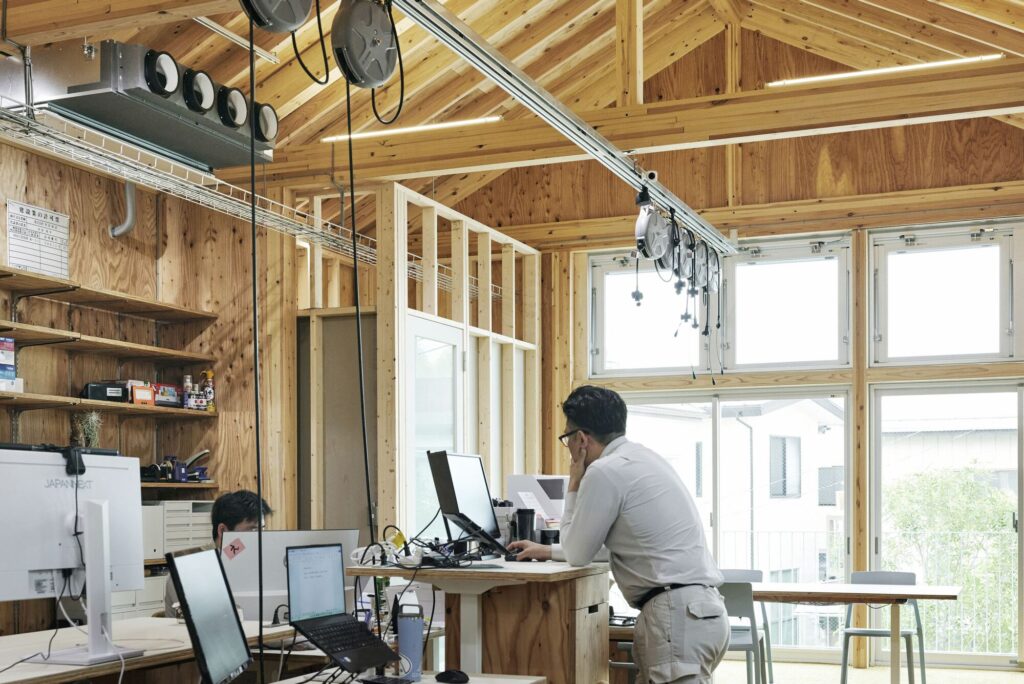 Office workers at standing desks inside a wooden-structured room with exposed gabled roof beams and natural lighting.