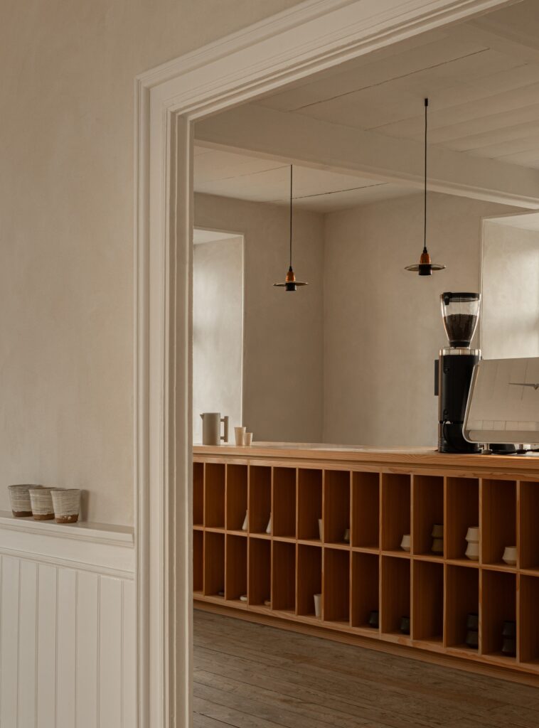A view of a wooden service counter with integrated shelving for ceramics and minimalist black pendant lights hanging from a white ceiling.