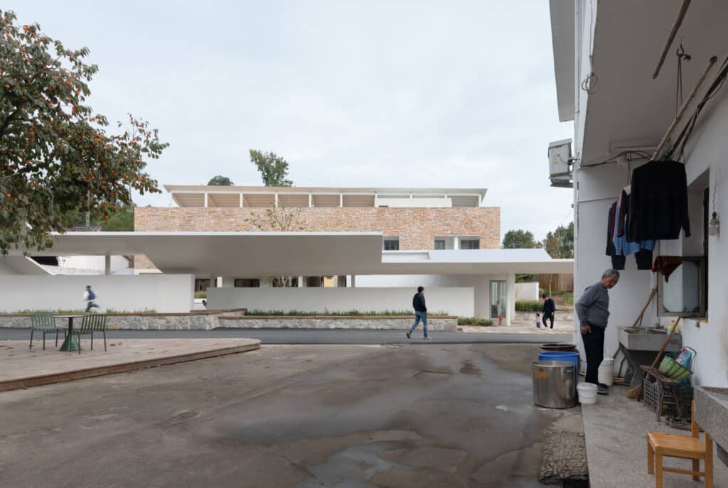 Wide shot showing a village street with residents and the modern pavilion of Qingyi Lodge in the background.