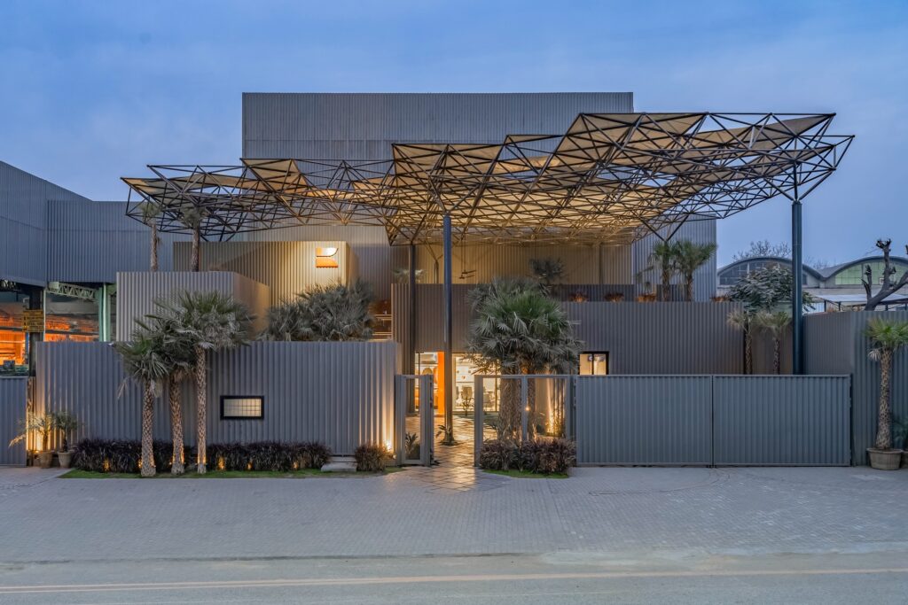A night view of the Karachi Steel warehouse featuring a dynamic space frame structure over the entrance and a grey corrugated facade with integrated greenery.