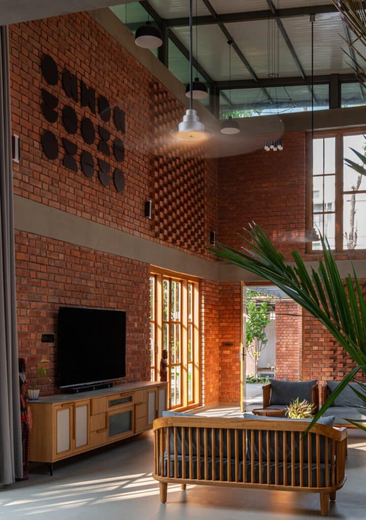 Double-height living area with brick walls, wooden furniture, a large TV unit, and high windows letting in natural sunlight.