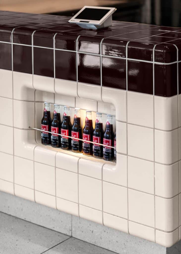 A close-up of a cream-tiled service counter featuring a recessed display shelf with glass soda bottles under warm accent lighting at North of Brooklyn Pizzeria.