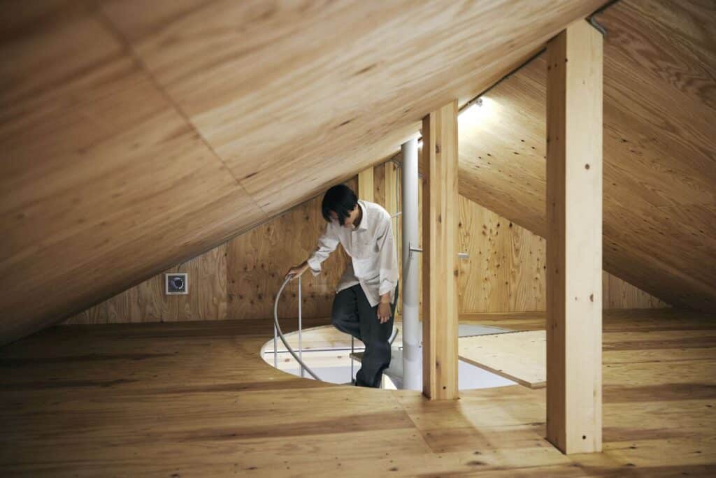 A person descending a spiral staircase from a spacious wooden loft area under a slanted plywood ceiling.
