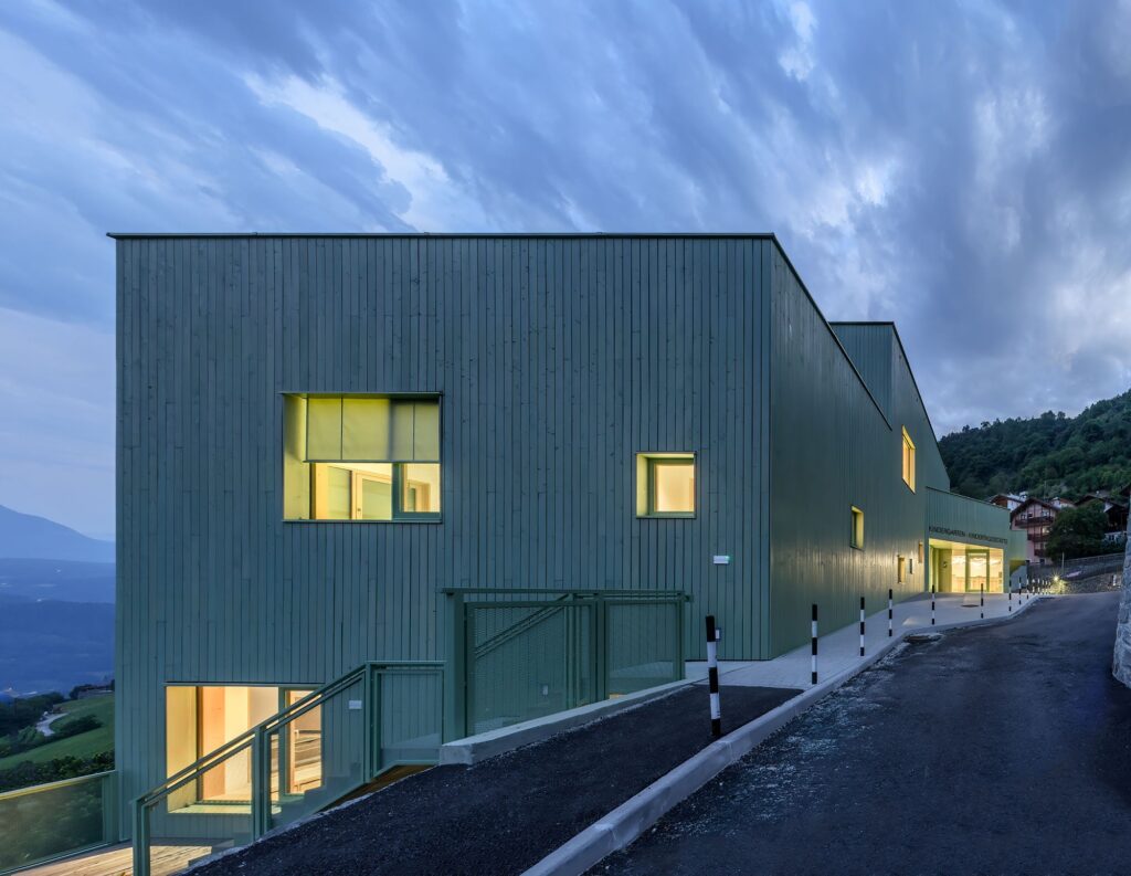 Evening view of the Barbiano kindergarten's green timber exterior with warm light shining through square windows under a blue twilight sky.