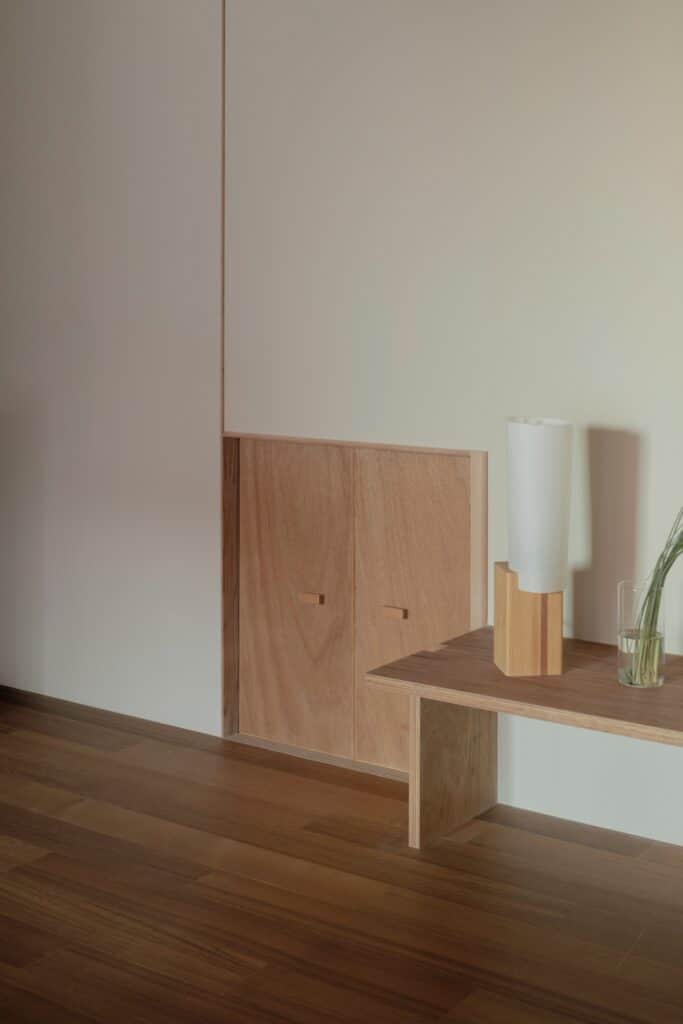 A minimalist white wall with a small wooden storage door and a simple wooden bench.