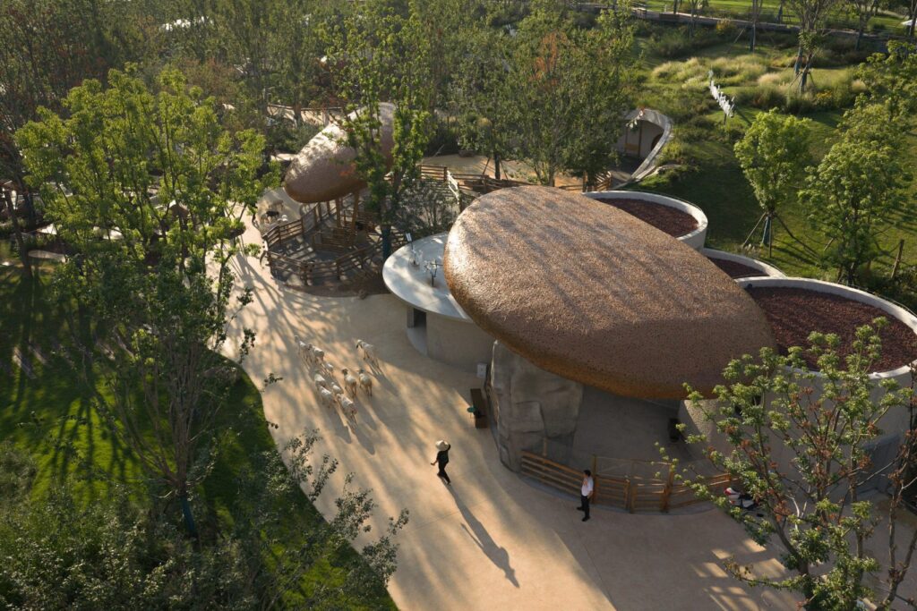 An aerial view of an organic-style farm landscape featuring large, pebble-shaped textured roofs over animal enclosures, with a small flock of sheep being led down a winding path by a person in a sun hat.